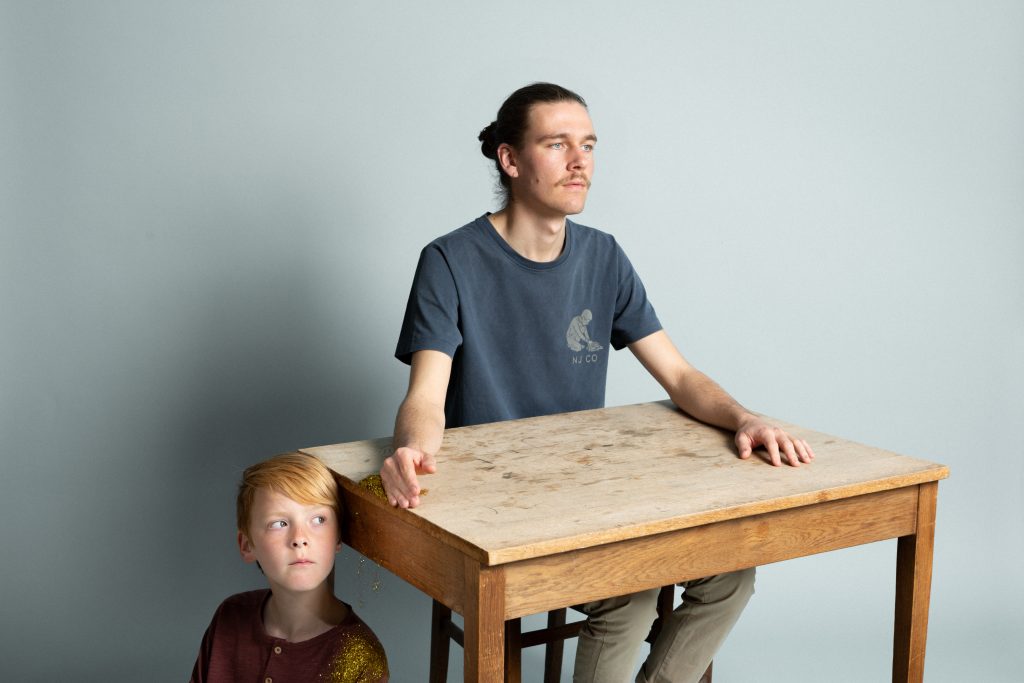 Een persoon zit achter een tafel en kijkt voor zich uit.
Een kind leunt met zijn hoofd tegen de tafel en kijkt in dezelfde richting